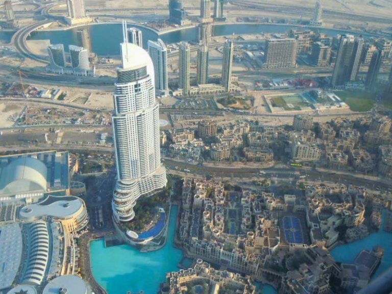 Aerial view from Burj Khalifa observation deck looking down at Dubai's skyscrapers, turquoise fountains, and surrounding city with construction areas visible in the distance.