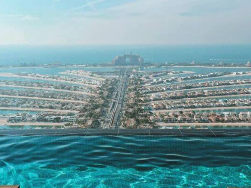 A view of Palm Jumeirah from the Aura Sky Pool View of Palm Jumeirah in Dubai from Aura Sky Pool with turquoise infinity pool water in the foreground and the palm-shaped island stretching into the sea with Atlantis hotel visible in the distance.