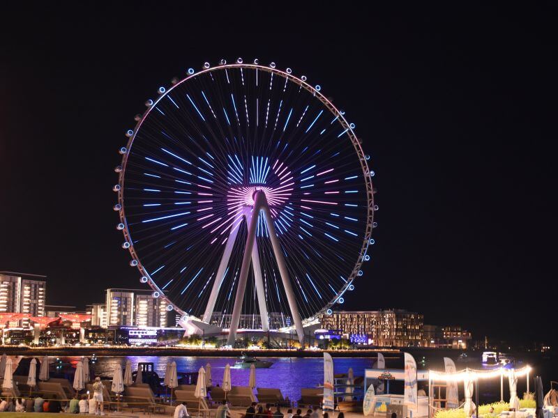 Ain Dubai observation wheel lit up with blue and pink lights at night, viewed from JBR beach with waterfront seating in the foreground.