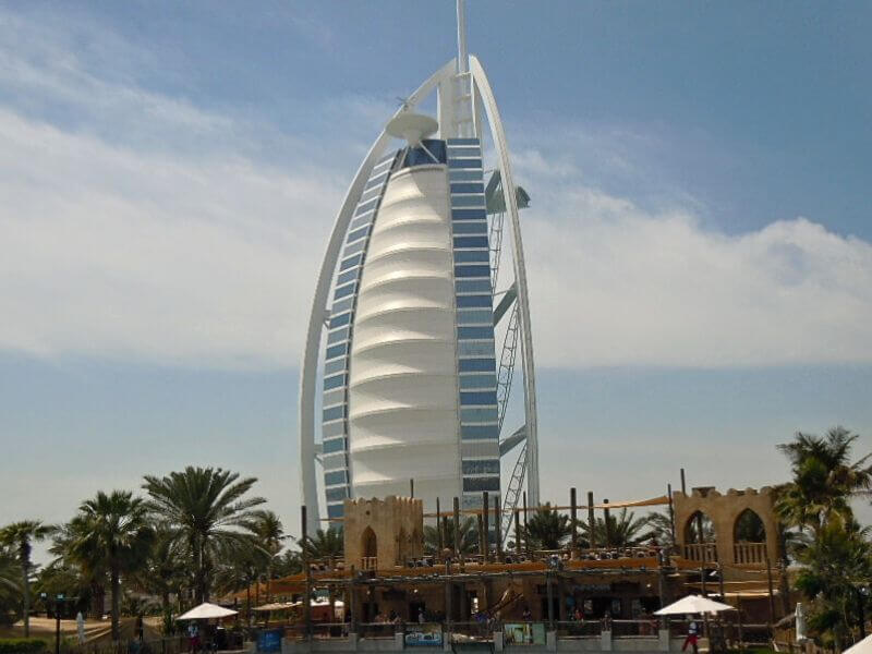 Burj Al Arab, Dubai Burj Al Arab hotel in Dubai with its white sail-shaped design, palm trees and beachfront buildings in the foreground under a blue sky.