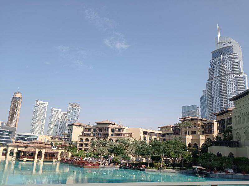 Downtown Dubai, Souk Al Bahar Souk Al Bahar shopping complex in Downtown Dubai with turquoise Burj Lake in front, palm trees, and tall skyscrapers in the background under a blue sky.