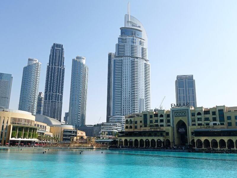 Downtown Dubai Downtown Dubai skyline with Burj Lake in the foreground, with calm blue water reflecting nearby buildings. Souk Al Bahar sits along the waterfront while tall modern towers rise behind it near the Dubai Mall. The image shows a bright, clear day and a lively city area by the water.