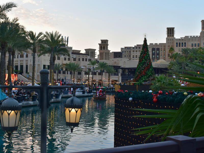 Dubai Madinat Jumeirah at Christmas Madinat Jumeirah in Dubai decorated for Christmas with a large lit tree beside the water. People walk along the waterfront while lights reflect on the canal and traditional buildings stand in the background.