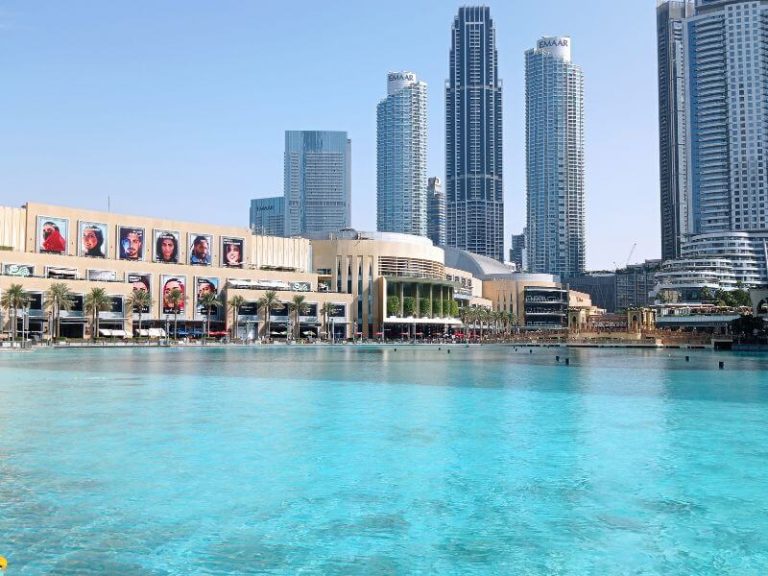 Dubai Mall with turquoise Burj Lake in front and tall skyscrapers in the background under a blue sky.