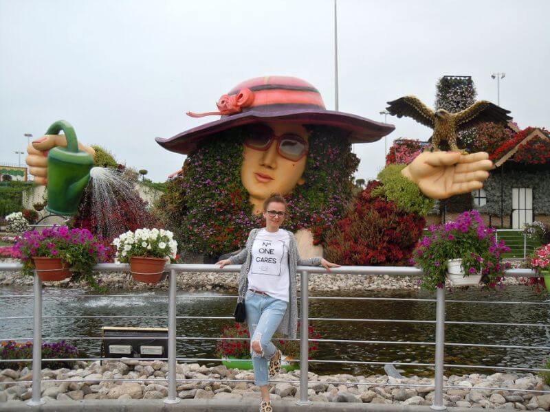 Dubai Miracle Garden Woman standing in front of a large flower sculpture of a lady with a hat and watering can at Dubai Miracle Garden with colorful flowers and plants.