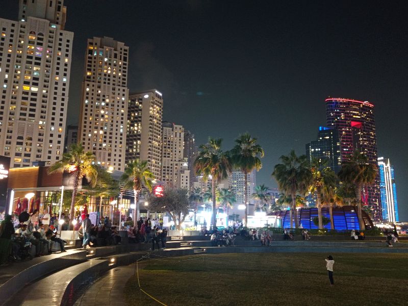 JBR (Jumeirah Beach Residence) in Dubai at night with people gathered on the lawn, palm trees, lit residential towers, and waterfront area with colorful lights.