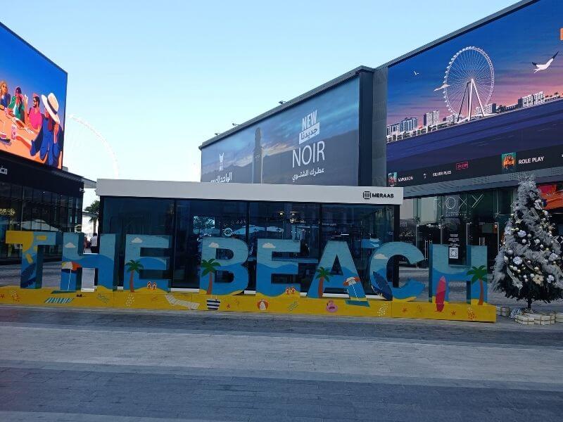 Jumeirah Beach Residence, Dubai Outdoor view of Jumeirah Beach Residence with a large The Beach sign in front of shops and walkways. Digital screens and buildings line the open area with a clear sky above. The scene shows a popular spot where people walk, shop, and enjoy time near the beach.