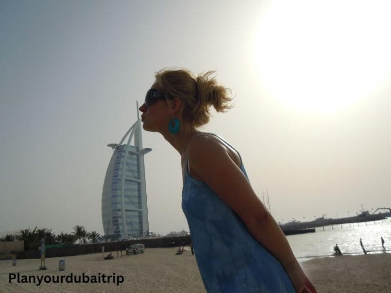 Woman playfully posing as if kissing the Burj Al Arab hotel in Dubai, with the iconic sail-shaped building visible in the background on the beach.