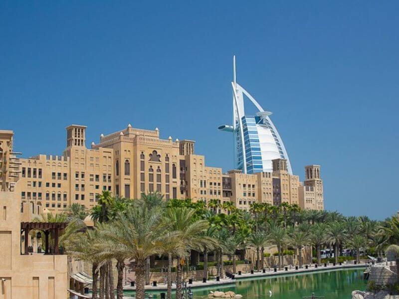 Madinat Jumeirah, Dubai Madinat Jumeirah in Dubai with traditional Arabic buildings, palm trees, green waterway, and Burj Al Arab hotel visible in the background under a blue sky.