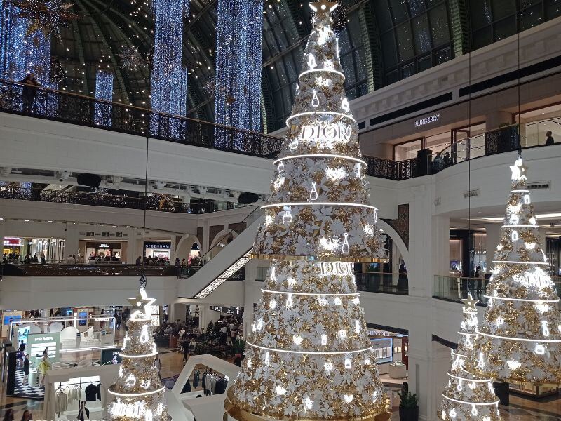 Mall of the Emirates in December Inside Mall of the Emirates in Dubai during December, large Christmas trees decorated with lights and ornaments fill the space. Shoppers walk around stores like Prada and Burberry while festive lights hang from the ceiling.