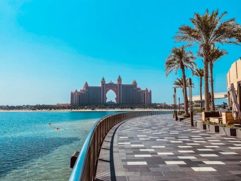 Palm Jumierah, Dubai Waterfront view of Palm Jumeirah with a wide walking path lined by palm trees beside the sea. The Atlantis hotel stands in the distance across the water, facing the curved shoreline. The image shows a calm place where people walk and enjoy views of the ocean in Dubai.