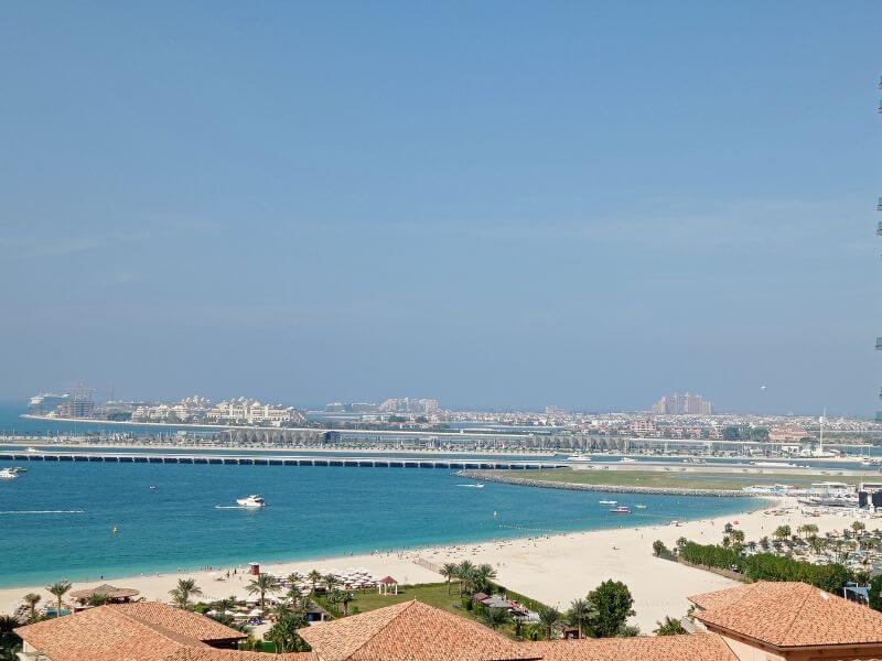 Sea and beach view in Dubai Beach and turquoise sea view in Dubai with white sand, palm trees, hotels along the coast, and Palm Jumeirah visible in the distance under a blue sky.