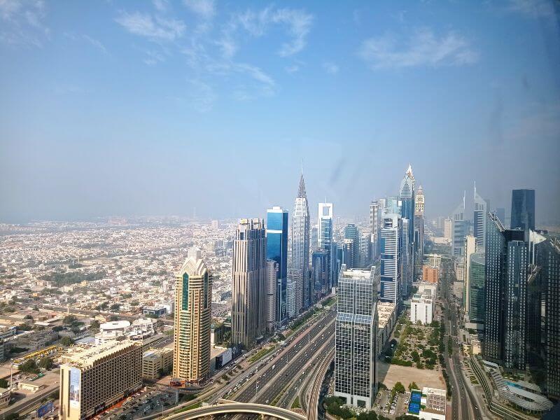 Aerial view from Sky Views Dubai showing the tall skyscrapers of Sheikh Zayed Road with highways running through the city and residential neighborhoods spreading into the distance under a blue sky.