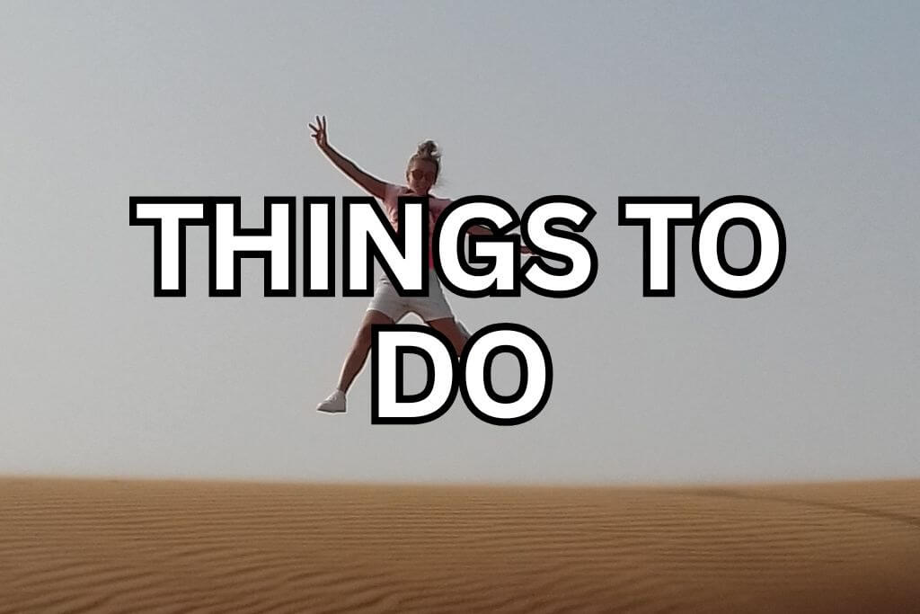 Person jumping in the Dubai desert with sand dunes stretching out below and a clear sky above. Large text over the image reads THINGS TO DO. The image suggests fun activities and experiences to enjoy while visiting the Dubai desert.