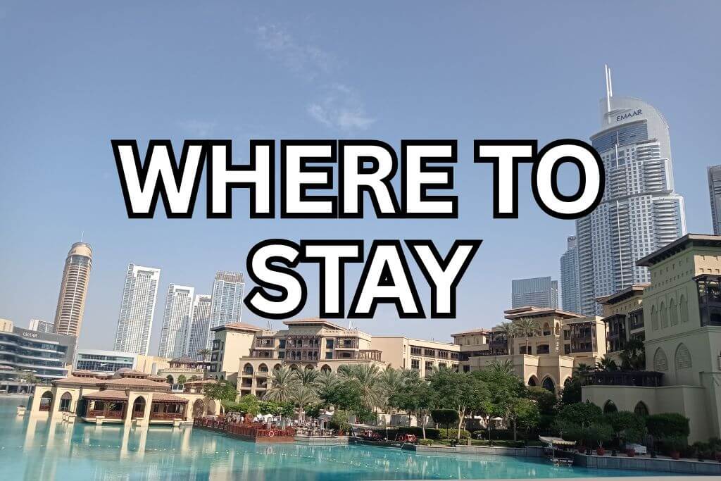 City view of Downtown Dubai with Souk Al Bahar with calm water in the foreground and modern skyscrapers in the background. Large text over the image reads WHERE TO STAY. The image gives a clear sense of the area and highlights it as a place to stay while visiting Dubai.