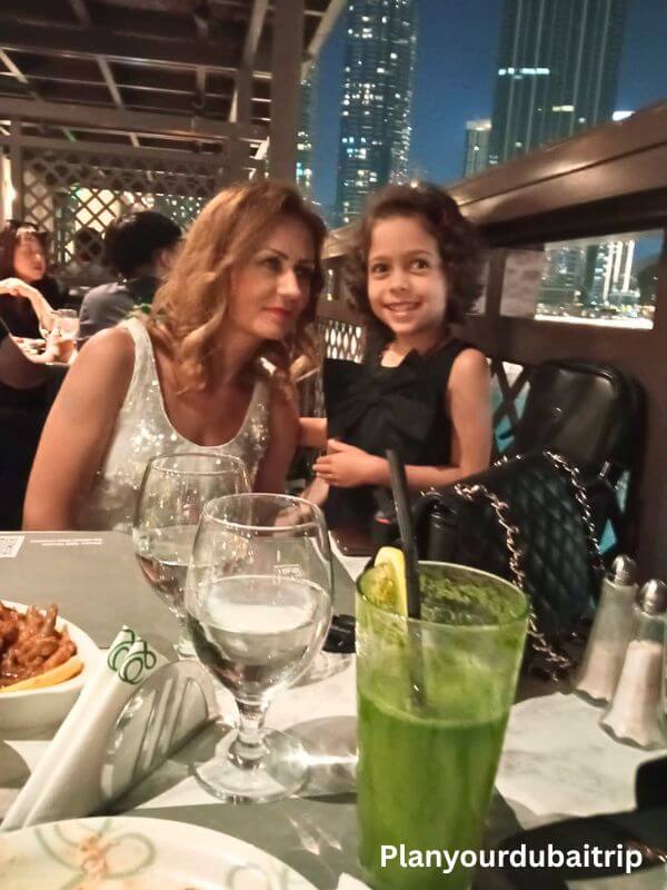 Abd El Wahab Souk Al Bahar, Dubai A woman and a young girl smiling at the dinner table at Abd El Wahab Restaurant in Souk Al Bahar, Downtown Dubai, with a fresh green drink and the city skyline lit up at night in the background.