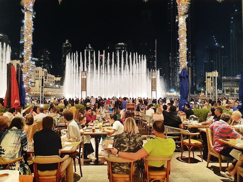 Outdoor seating at Carluccio's Restaurant in Dubai Mall with diners watching the Dubai Fountain show at night with palm trees wrapped in lights.