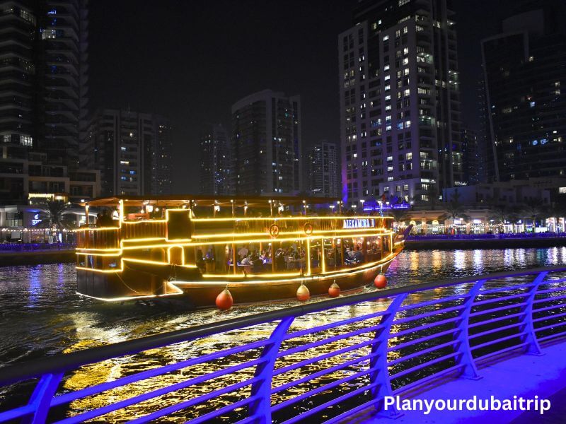 Dhow cruise in Dubai Marina Traditional dhow cruise boat lit up with golden lights sailing through Dubai Marina at night with tall buildings and waterfront restaurants in the background.