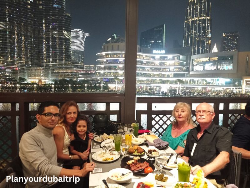 Dinner at Abd El Wahab Restaurant, Dubai A group of people enjoying dinner at Abd El Wahab Restaurant in Souk Al Bahar, Downtown Dubai, with a night view of the Dubai Mall and city skyline in the background.