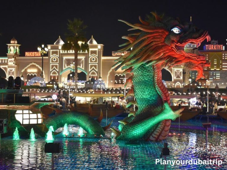 Large colorful dragon sculpture lit up with red and green lights in the water at Dubai Global Village with country pavilions in the background at night.