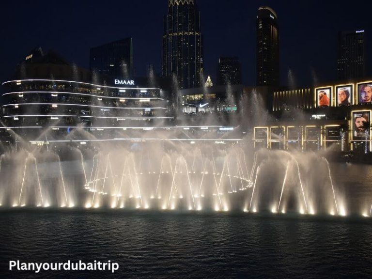 Dubai Fountain show at night with water jets lit up and dancing in front of Dubai Mall and tall buildings.