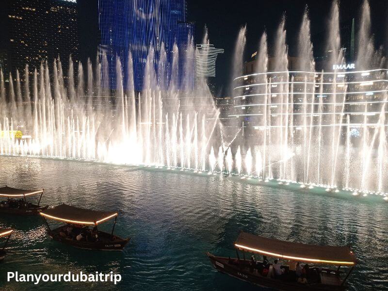 Dubai Fountain Show The Dubai Fountain show at night with water jets lit up against the city skyline, seen from Abd El Wahab Restaurant in Souk Al Bahar, with traditional wooden abra boats floating on the water in the foreground.