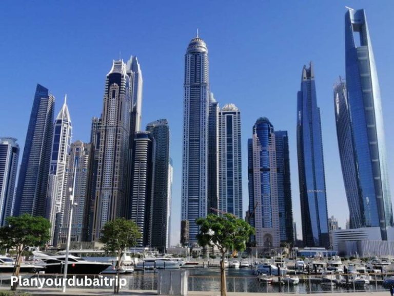 Dubai Marina skyline on a sunny day with tall skyscrapers, boats docked in the marina, and palm trees along the waterfront.