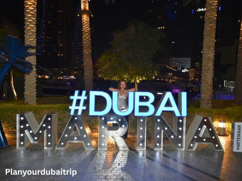 Dubai Marina sign Woman posing behind the lit-up Dubai Marina sign at night with palm trees and tall buildings in the background.