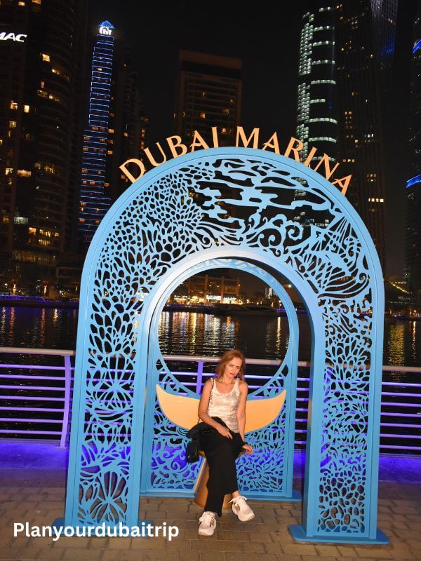 Dubai Marina Woman sitting in a blue Dubai Marina arch at night with the marina waterfront and tall buildings lit up in the background.