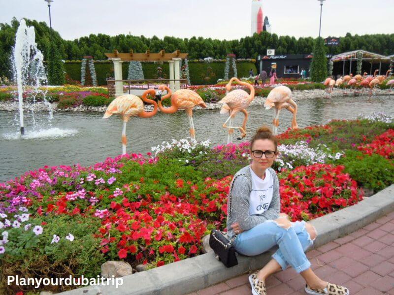 A woman sitting next to a flower bed at Dubai Miracle Garden, with flamingo sculptures in a pond and water fountains in the background.