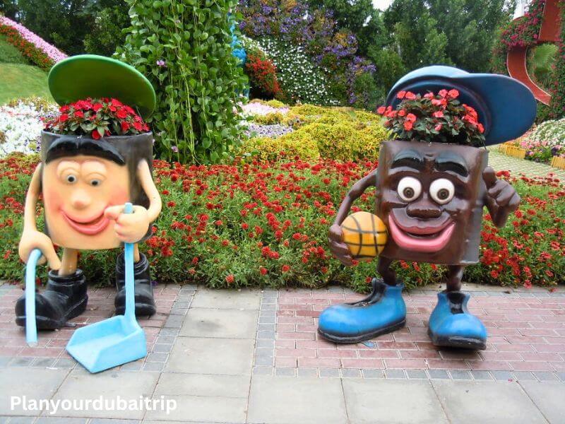 Two fun cartoon character sculptures with flowers growing out of their heads at Dubai Miracle Garden, surrounded by colorful flower beds.