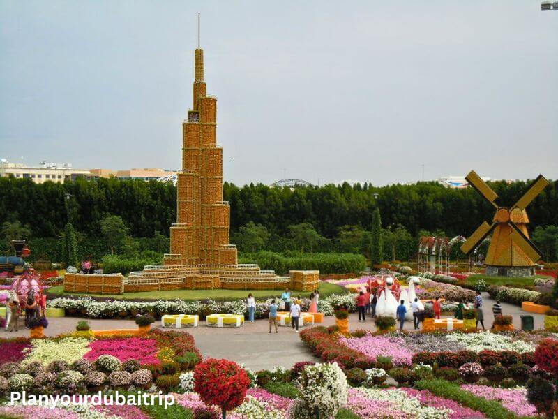 A flower replica of the Burj Khalifa surrounded by colorful flower beds and visitors walking around Dubai Miracle Garden, with a flower windmill in the background.