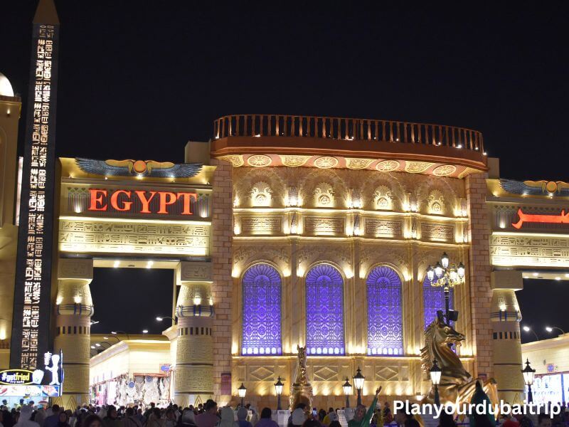 Egypt Pavilion in Dubai Global Village Egypt pavilion at Dubai Global Village lit up at night with Egyptian designs, purple lit arches, and a statue in front.