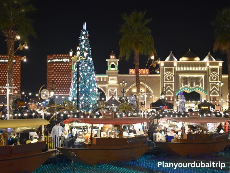 Floating market in Dubai Global Village Floating market with wooden boats selling food at Dubai Global Village with a large Christmas tree and lit-up pavilions in the background at night.