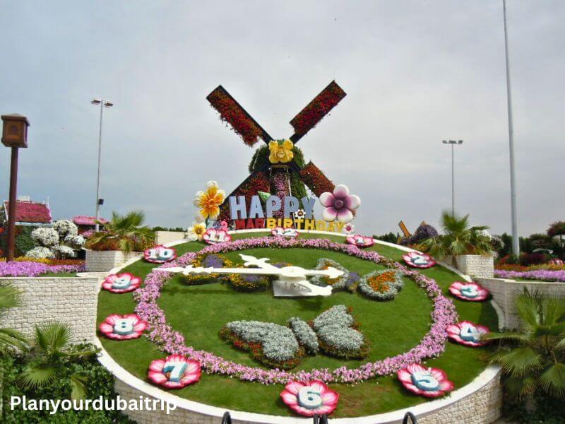 A large flower clock with colorful blooms at Dubai Miracle Garden, featuring a windmill covered in flowers and a Happy Birthday sign in the background.
