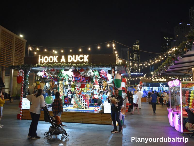 Fun for kids in JBR Dubai Hook a Duck game booth decorated with holiday lights and prizes at the kids area in JBR Dubai with families walking around at night.