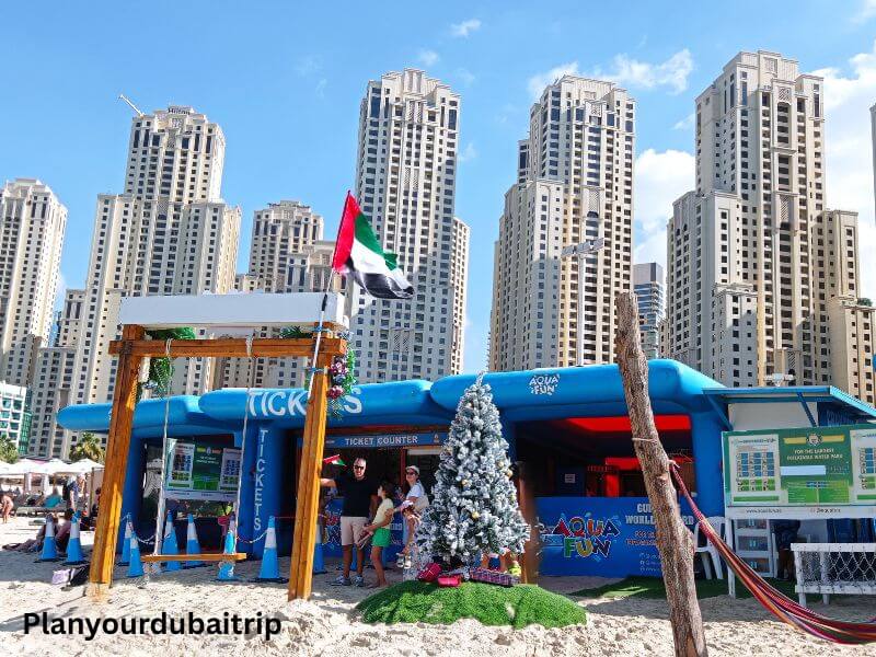 JBR Beach Dubai JBR Beach in Dubai with a blue water sports booth, UAE flag, small Christmas tree, and tall residential buildings in the background.
