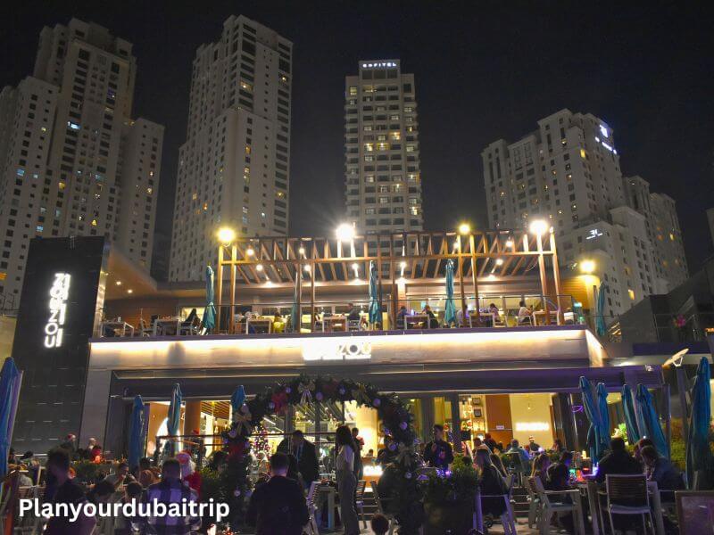 Restaurant in JBR Dubai Two-story restaurant at JBR Dubai lit up at night with diners on the rooftop terrace and people gathered below, surrounded by tall buildings.