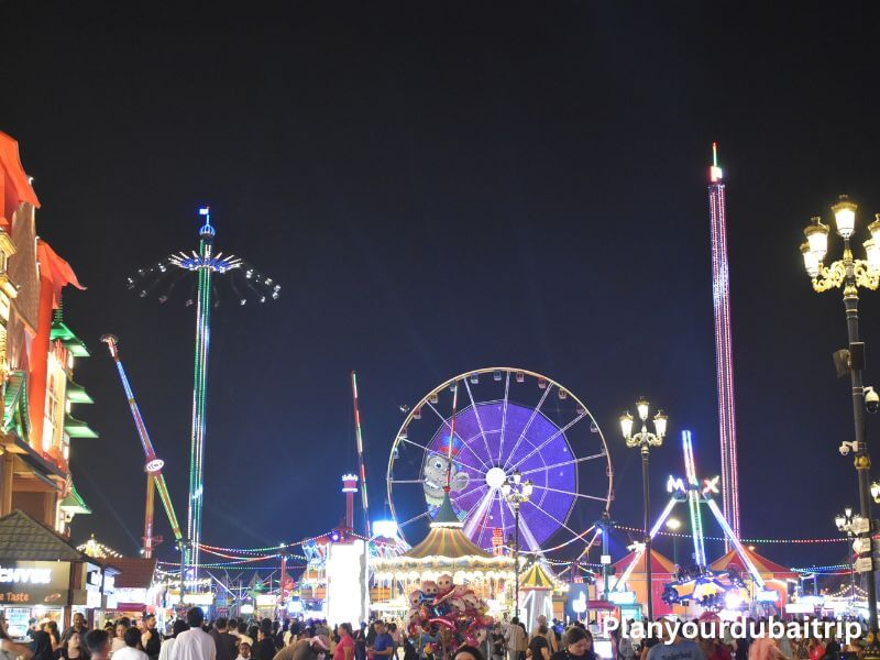 Rides in Dubai Global Village Amusement rides lit up at night at Dubai Global Village with a Ferris wheel, swings, and other colorful rides with crowds of people walking around.