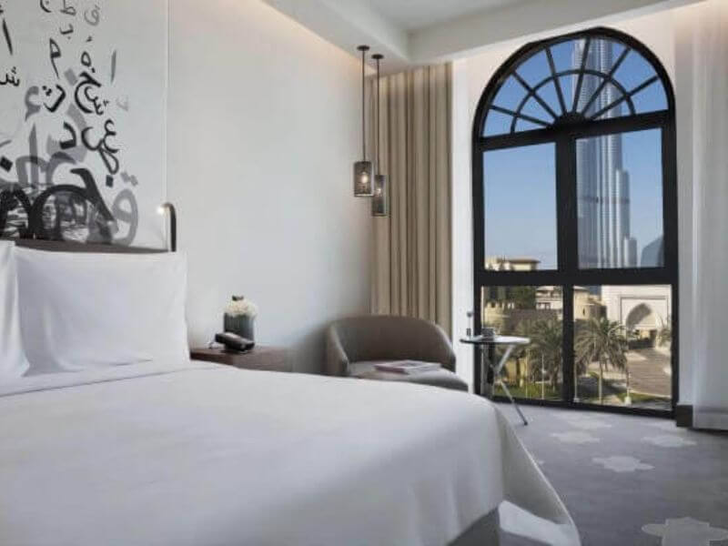 Guest room at The Heritage Hotel, Autograph Collection in Dubai with a white bed, arched window with Burj Khalifa view, and Arabic wall art.