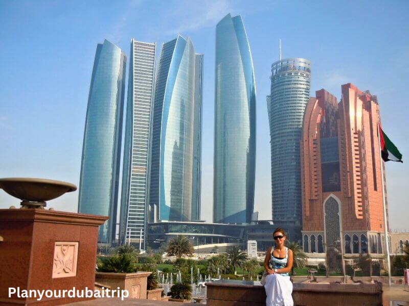 A woman posing in front of the Etihad Towers and the Emirates Palace Hotel in Abu Dhabi on a sunny day, with the UAE flag flying in the background.