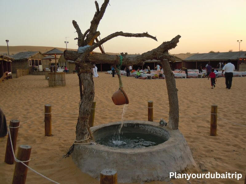 A traditional water well in the middle of a desert camp in Dubai, with sand dunes, straw huts, and visitors walking around in the background at sunset.