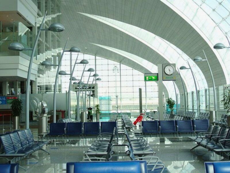 Dubai Airport A quiet departure gate area at Dubai International Airport with rows of blue seats, a curved glass ceiling, and a clock on the wall.