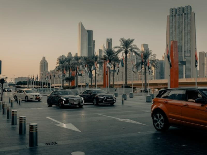 Dubai roads Cars driving on a wide road in Dubai at dusk, with palm trees, UAE flags, and tall buildings visible in the background.