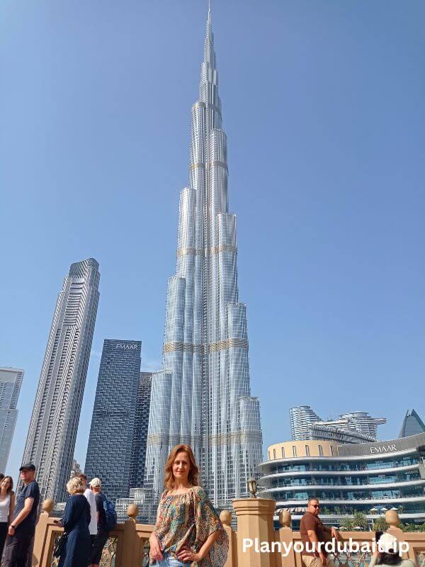 A woman posing in front of the Burj Khalifa on a sunny day in Downtown Dubai, with other tall buildings and the Dubai Mall visible in the background.
