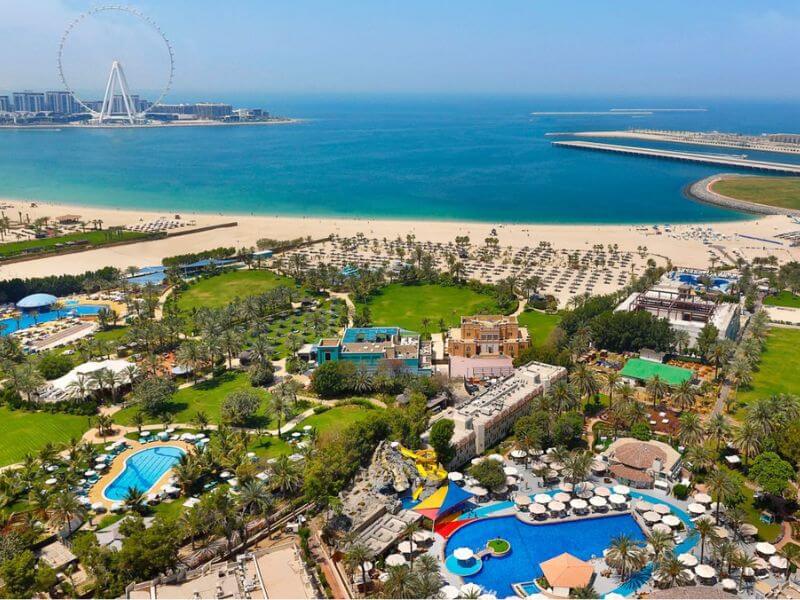 A top view of Al Habtoor Grand Resort Autograph Collection in Dubai, showing its pools, water slides, green gardens and palm trees, with a wide sandy beach, the blue sea and the Ain Dubai Ferris wheel visible in the background.