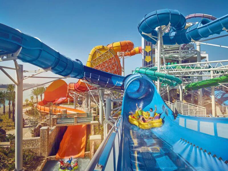 A group of people having fun on a yellow rubber ring going down a big water slide at Aquaventure Water Park in Atlantis The Palm hotel in Dubai, surrounded by colorful water slides on a sunny day.