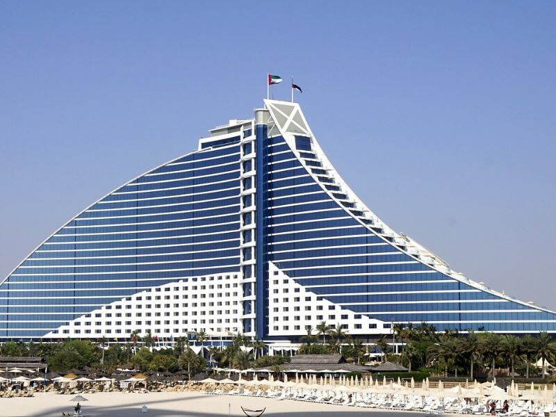 A view of the Jumeirah Beach Hotel in Dubai from the beach, showing its wave-shaped building with UAE flags on top, palm trees, and rows of sun loungers on the white sandy beach in front.