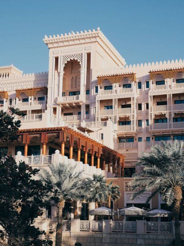 A close up view of the Jumeirah Dar Al Masyaf Hotel in Dubai, showing its white and gold Arabic style building with detailed archways, balconies, and palm trees in front.