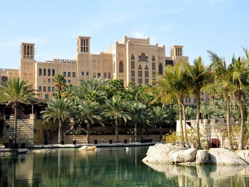 A view of the Jumeirah Mina Al Salam Hotel in Dubai from the waterway, showing its traditional Arabic style building surrounded by palm trees reflecting in the calm water.
